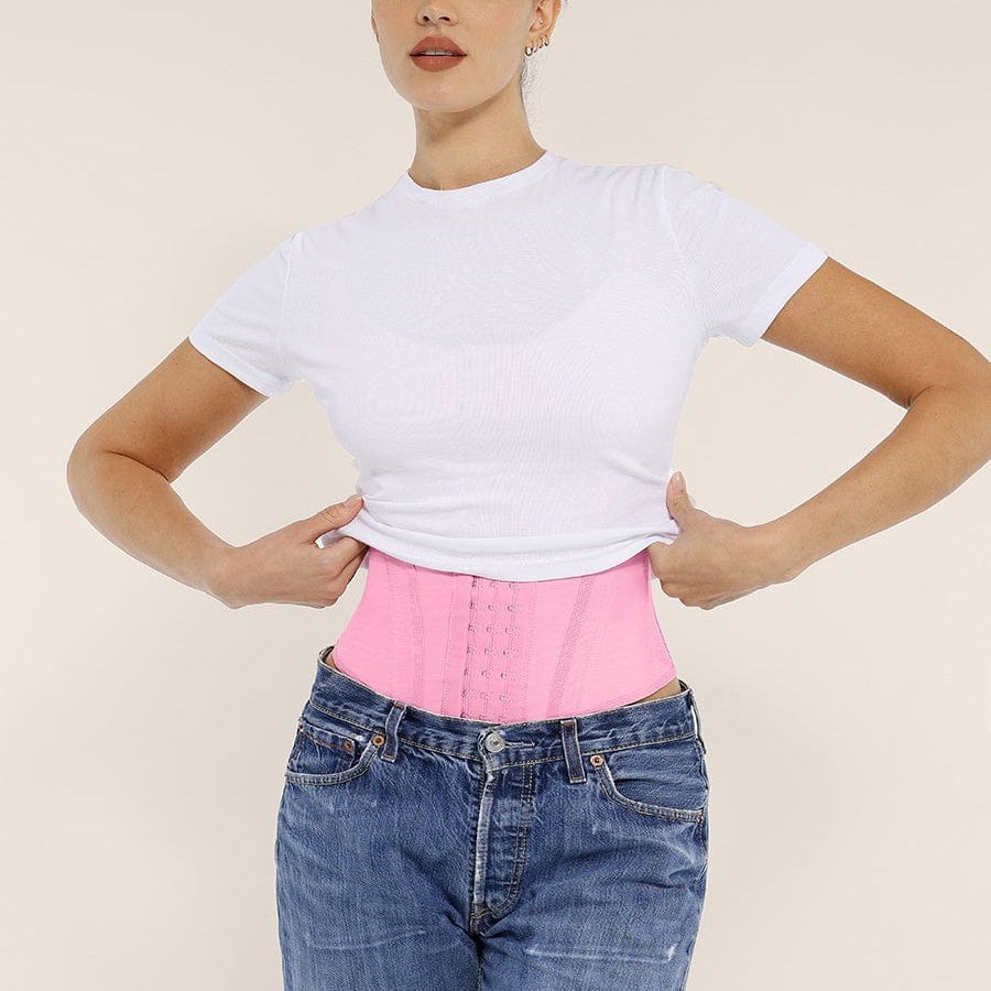 Woman wearing a pink waist trainer with a white shirt and blue jeans on a beige background